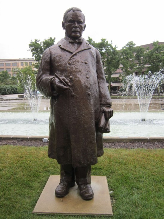 A statue of a lawyer with a receding hairline, a mustache, a trenchcoat, a clutch of legal papers and a cigar.