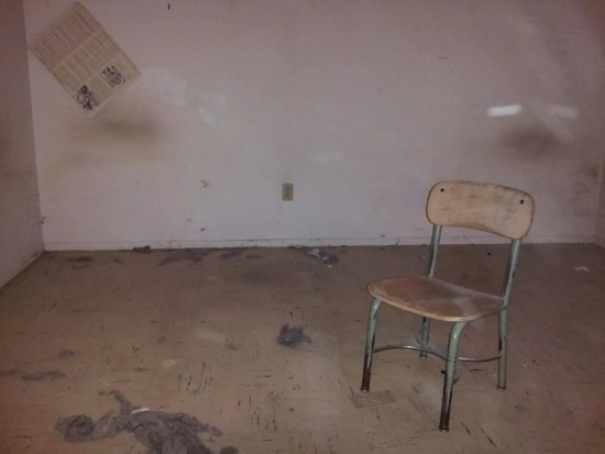 old wooden and metal school chair in the middle of an empty apartment with dust bunnies all over the floor and a taped poster drooping off a wall.