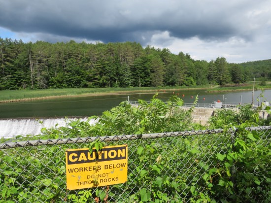 Large ponds that flows into the dam. A yellow sign reads "CAUTION: WORKERS BELOW, DO NOT THROW ROCKS". More practical advice.