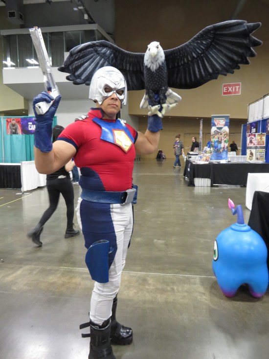 Cosplayer Peacemaker with an Eagly puppet on his arm.