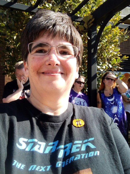 Anne takes a selfie in a Trek shirt. I'm at left over her shoulder, staring at my phone and holding my chin in my hand in a weird way that makes it look like I'm picking my nose.