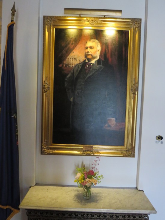 Painting: portly gentleman with muttonchops, White House in the distance over his shoulder. Below the painting is a vase of flowers on an otherwise empty table.