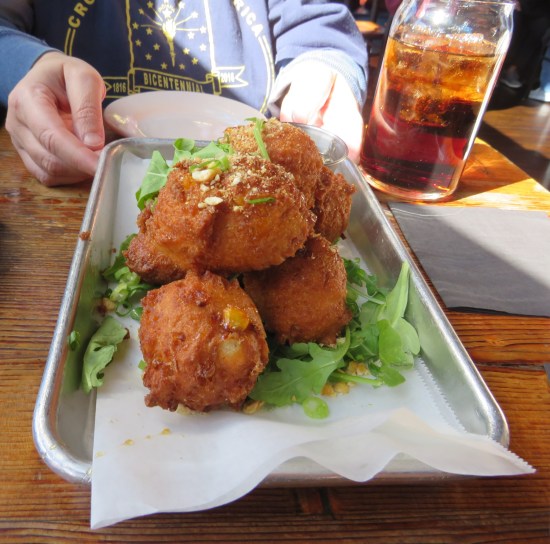 Ball-shaped corn fritters on a rectangular white plate. Behind them, Anne is wearing her Indiana Bicentennial shirt.