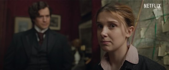 Millie Bobby Brown as Enola Holmes, perplexed at the camera while Henry Cavill's Sherlock stands out-of-focus behind her.