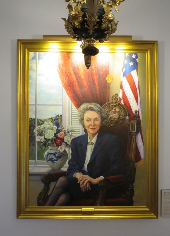 Painting: shrewdly smiling businesswoman sitting by a window, a flag, and a vase of flowers.
