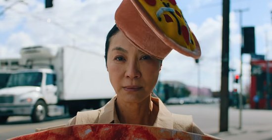 Michelle Yeoh in a beige suit wearing a hat shaped like a pizza, cocked to one side with the nonchalance of someone who ALWAYS wears pizza hats.