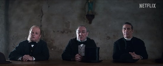 Three old men sitting in black robes at a table and beseeching an offscreen nurse for help with their small town's weird problem.