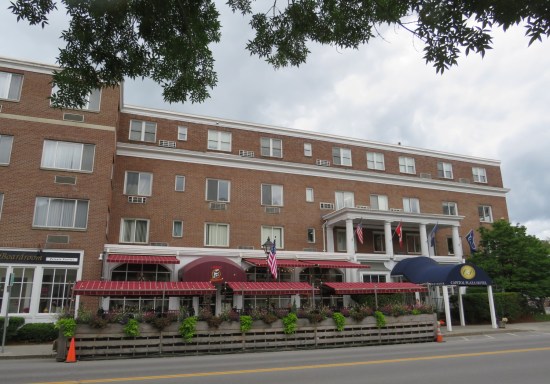 A four-story brick hotel with a restaurant on the ground floor, with red awnings and no customers seated at the outside tables.