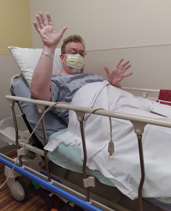 Me in a hospital bed, masked and hooked to an IV and doing jazz hands anyway.