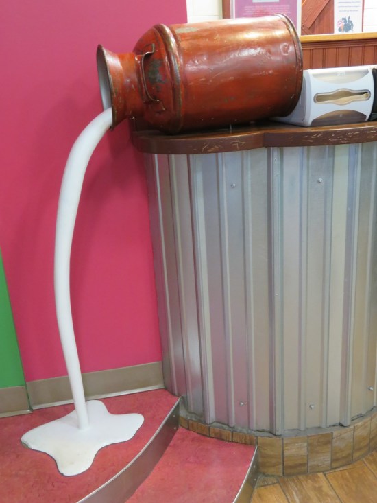 In statue form, a jug of milk tipped over and pouring fake milk on the floor.