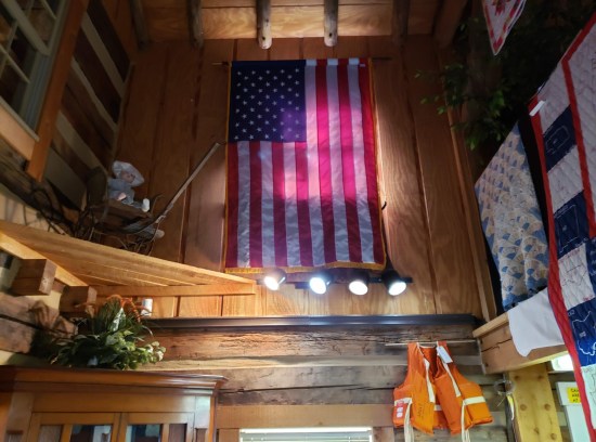Hanging near the ceiling is a sideways US flag, mostly in shadows with a lone weird light shining near its center. At left in the shadows is a creepy baby doll.