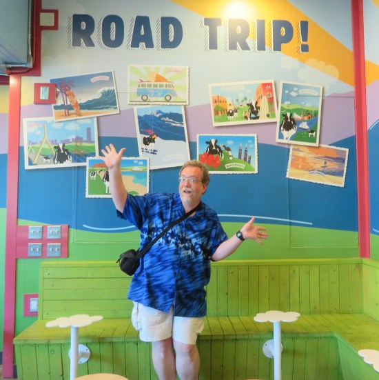 me doing jazz hands in front of a wall titled "ROAD TRIP!" Painted on the wall are photos of cartoon cows traveling to famous places around the world.