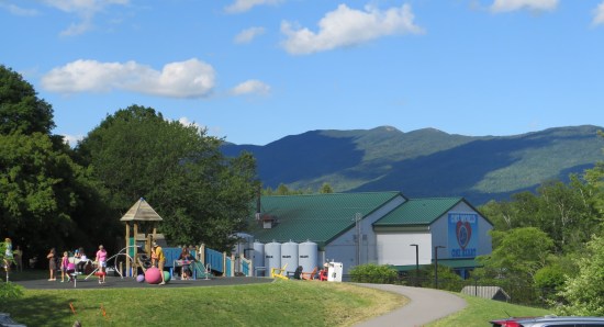 At left, a playground; at right, the factory in the distance with giant liquid tanks attached in back; on the horizon, mountains.