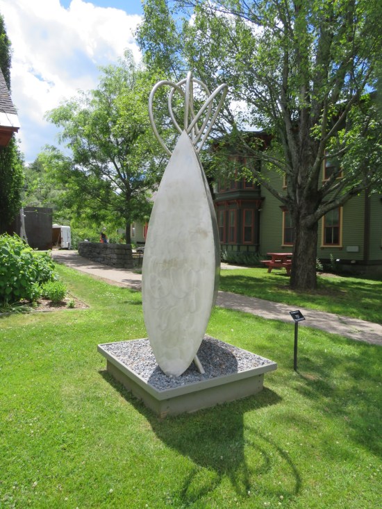 A steel sculpture that resembles an oblong leaf standing on one end, leaning a tad, with metal loops on top.