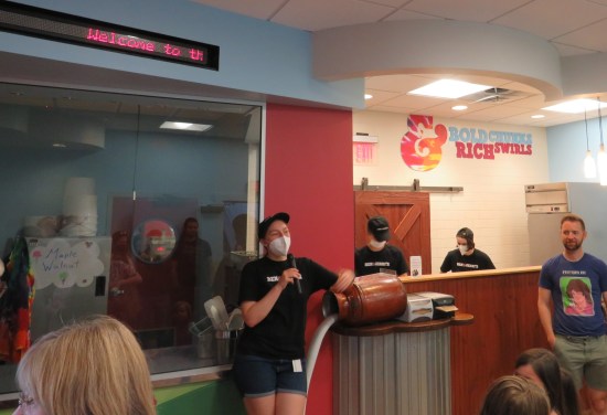 Three factory employees: a tour guide with a microphone in front of a window and two servers preparing to hand out free ice cream.