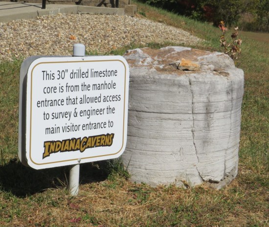 A limestone cylinder sitting outside with a sign: "This 30-inch limestone core is from the manhole entrance that allowed access to survey & engineer the main visitor entrance to Indiana Caverns."