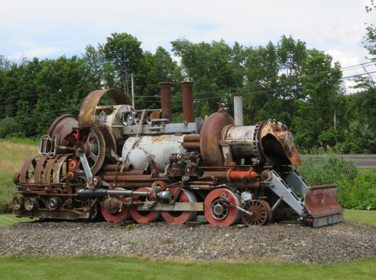 A nearly full-size train engine composed entirely of recycled gadgets and gizmos, sitting on a grassy field.