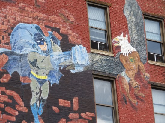 A mural painted on a brick wall depicting Batman busting through the wall. Flying next to him is a griffin with a superhero-stylized "G" on its chest.