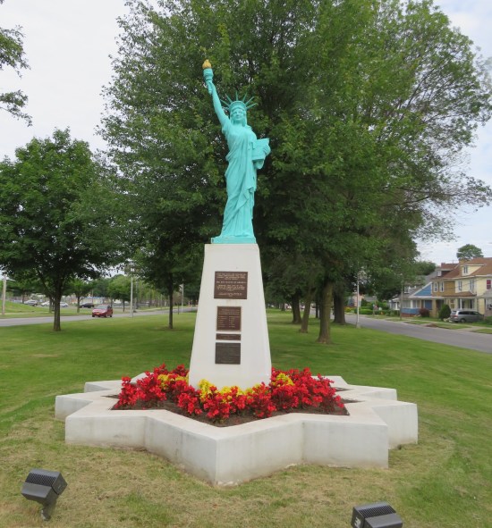 A tiny Statue of Liberty atop a pedestal surrounded by red flowers inside a starburst-shaped cement-walled garden.