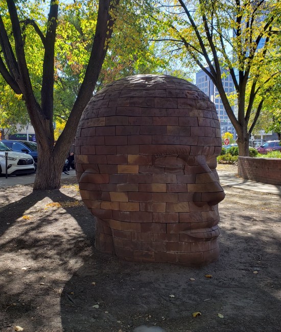 A giant human head made of bricks, surrounded by dirt but shaded by several nearby trees.