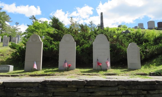 Four tombstones in a row, with small American flags stuck in the grass in front of them.