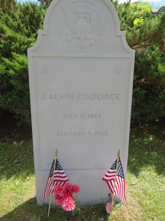 Calvin Coolidge's tombstone has two tiny flags and some pink flowers standing in front of it; evergreen bushes behind it.