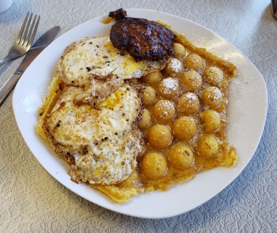 A cornbread waffle with bubble shapes poking out of the top. Eggs and sausage share the plate.