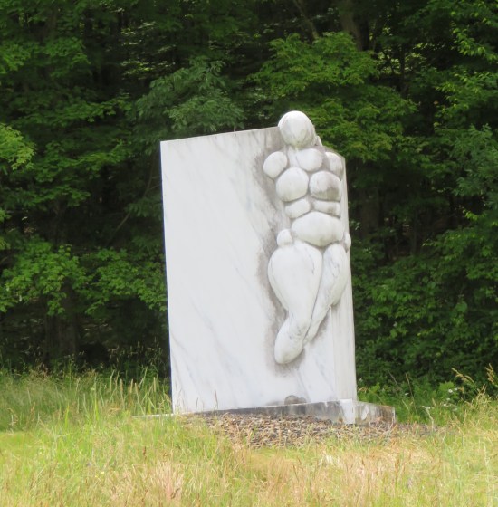 A 7-foot-tall marble block with a faceless, big-boned marble woman attached on one corner, a foot above the ground.