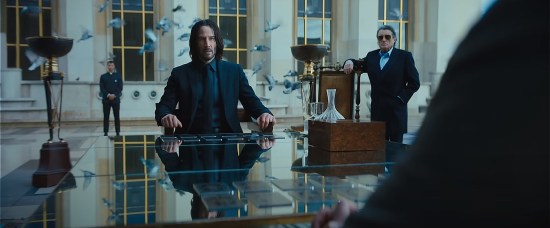 Keanu Reeves' John Wick sits outdoors at a shiny table with two rows of glass cards in front of him, and two identical rows in front of the unseen opponent seated across from him. Off to one side, Ian McShane in sunglasses looks sternly at the game. Blurry doves fly by in the background.