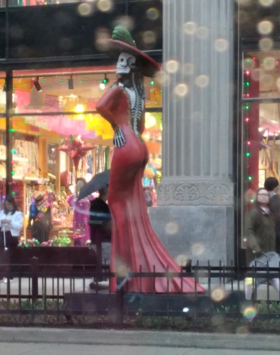 Statue of a lady skeleton in a red dress standing in front of upscale department stores.