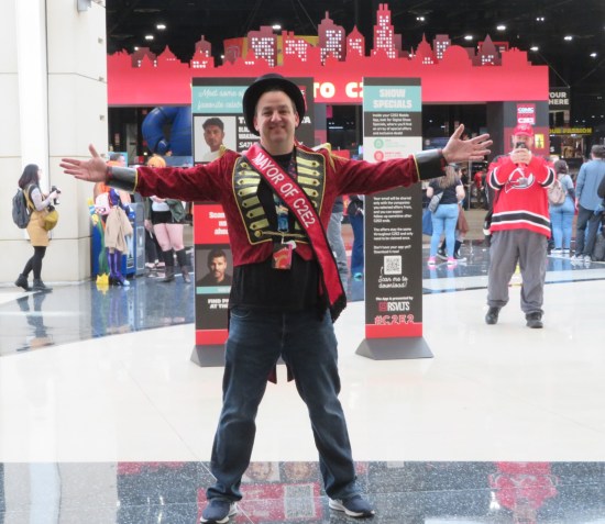 A happy dude in a red dress jacket, top hat and red "MAYOR OF C2E2" sash. His arms are widely welcoming as he stands in front of the exhibit hall gate.