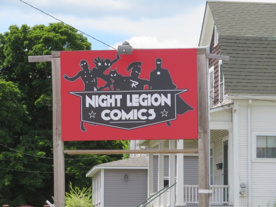 Red flag-shaped sign bearing Night Legion Comics' white-on-black medal-shaped logo. Behind the logo are silhouettes of five made-up superheroes.