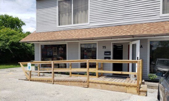 A ground-level comic shop in a 2-story building with a long, unpainted wooden wheelchair ramp leading up to the open front door.