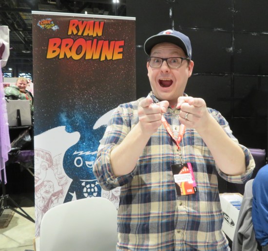 Ryan Browne happily pointing at you while standing in front of his table banner.