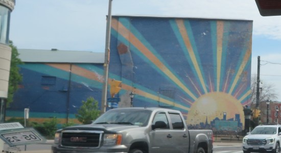 Mural: blue exterior wall on a building; at lower right is the sun, with large yellow rays emanating outward across the rest of the wall.