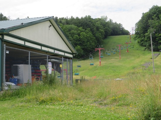 An entire empty ski lift suspended above a small, grassy hill. The cables run down toward a fenced building containing innertubes and other outdoor accoutrements.