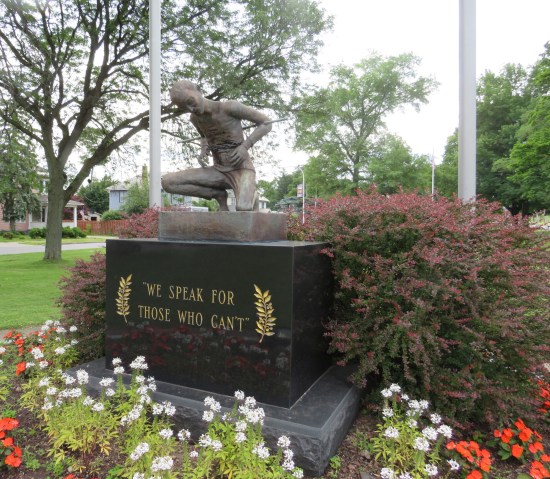 A statue of an emaciated P.O.W. on a rectangular pedestal surrounded by flowers. 