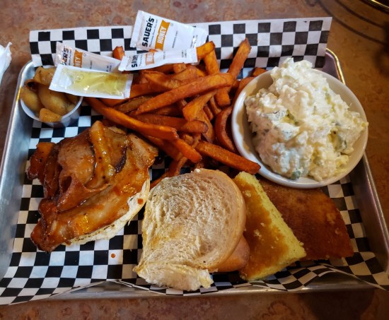 A metal tray containing a brisket sandwich, sweet potato fries, cole slaw, potato salad, AND warm apples.