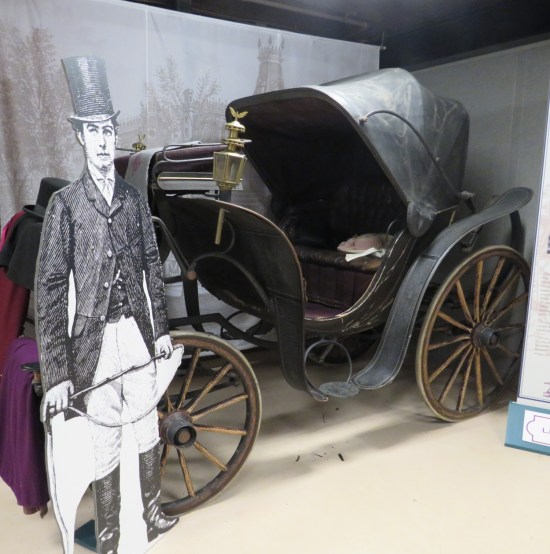 An old horse-drawn carriage parked in a basement, minus horses. It has oil lamps on either side of the driver's seat.