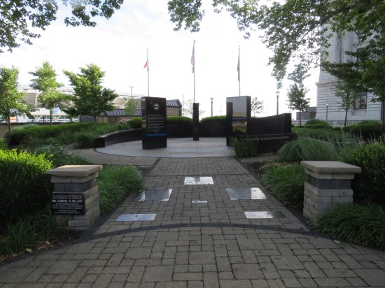 A memorial at the end of a brick path. Three flags, a sloping wall, some inscribed obsidian slabs.