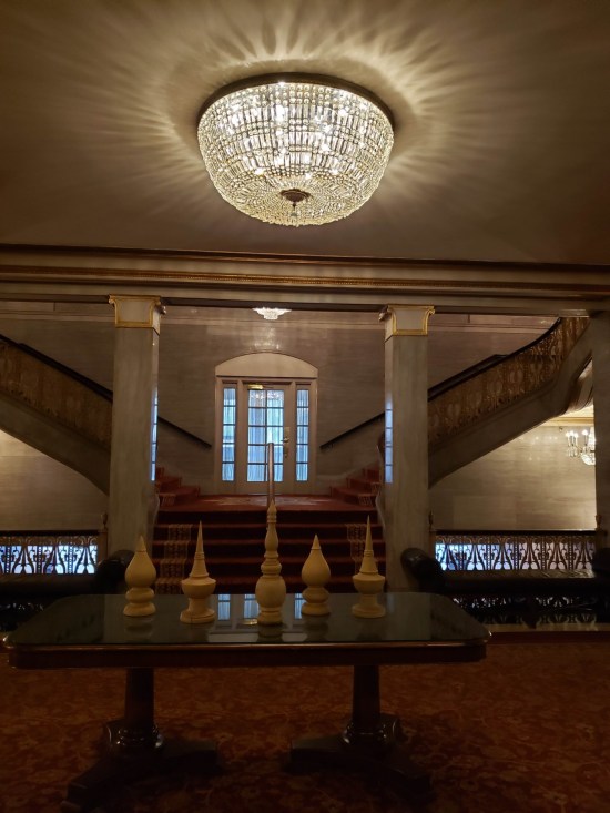 A grand hotel lobby with fancy domed ceiling light cover and a table with five weird wood-carved objects, each resembling mosques, chess pieces, or some other kinds of spires.