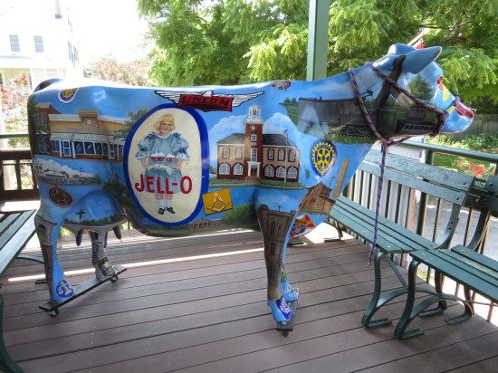 A cow statue on a balcony, painted mostly blue. Decorations include an ancient Jell-O ad with a tiny girl, and symbols of various small-town businesses nearby.