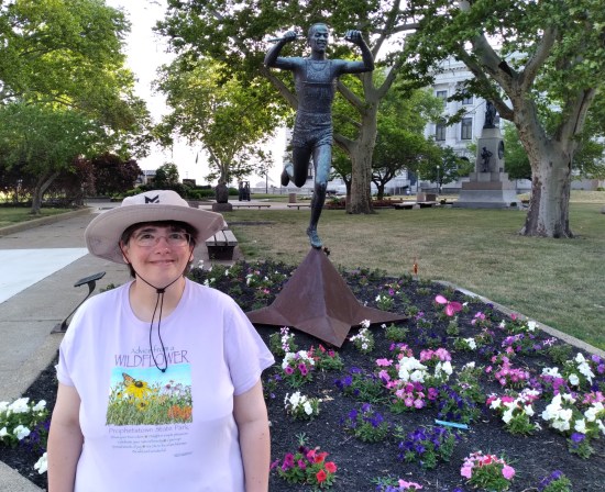 Anne smiling next to the Jesse Owens statue and a bed of pretty flowers.