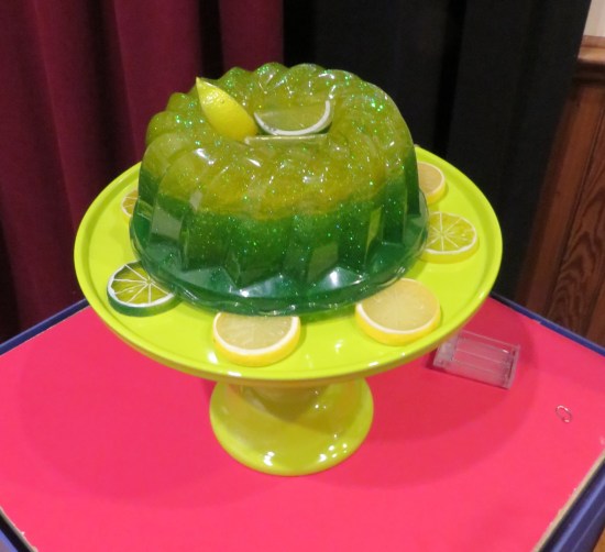A replica green Jell-O cake-sized dessert on a cake stand. Probably not made fresh that day.