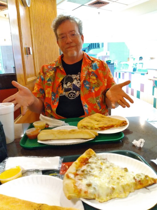 Me sitting at one end of a small table with two large slices of pizza on it, each on separate trays. One of them is closer to Anne the photographer.