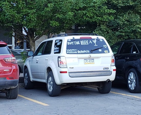 A parked silver van with "Buy the Bride a Drink!" written on the back window, along with someone's Venmo account name.