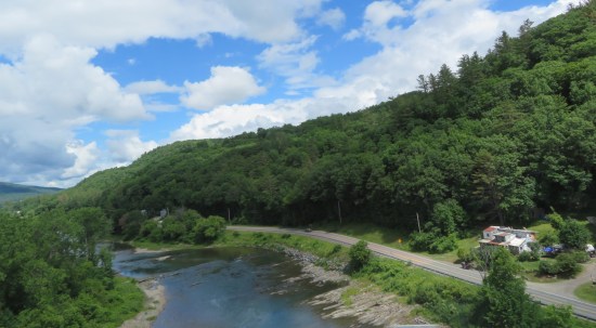 The deeply forested Green Mountains from afar on a brightly cloudy day. Someone's house is in the lower right corner; a waterway runs diagonally along the mountain bases.