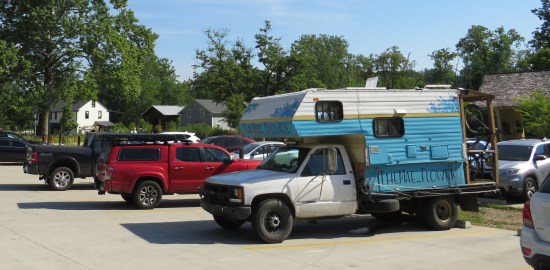 Three trucks in a parking lot, one of which is a camper truck with "THE MAE FLOWER" painted in large letters on the side.