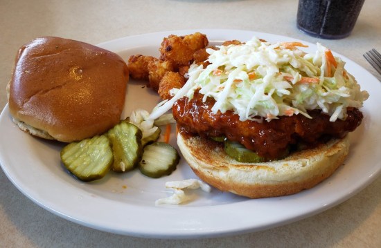 A breaded Chicken sandwich in hot sauce, topped with pickles and cole slaw. Behind it on the plate are some tater tots.