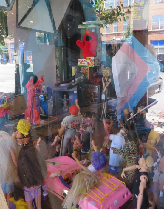 A pile of toys in a store window with an Elmo doll on top and other dolls standing around the base of his pyramid.